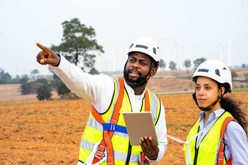 Two industrial engineers in hard hats discuss maintenance plans for rotating wind turbines.