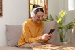 © amnaj - Cute Asian young woman wearing headphones sit on sofa and using smartphone to listen to music happy and relax listening to music at home.