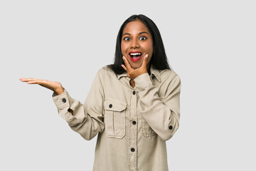 Wall Mural - Young Indian woman cut out isolated on white background holds copy space on a palm, keep hand over cheek. Amazed and delighted.