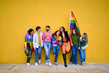 Portrait Of Group Cheerful Young Friends Standing On Wall Yellow. LGBT Community Smiling People On Gay Pride Day Enjoying Together And Showing Rainbow Flags. Generation Z And Parade Party Homosexual.
