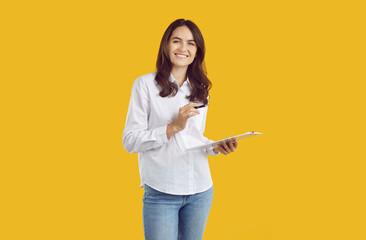 Portrait of happy young female secretary, office manager or business assistant with clipboard. Pretty woman in white shirt standing isolated on yellow background, holding clipboard and pen and smiling