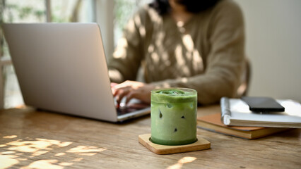 A glass of iced matcha green tea on a wooden table with a female sitting behind.