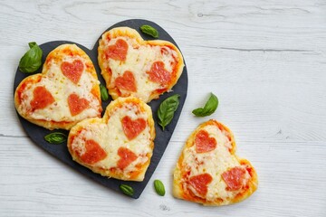 Wall Mural - Heart shaped mini pizzas with heart shaped pepperoni on heart shaped slate plate with white wood background.Creative art food idea for celebrate Valentine or Mother day.Top view.Copy space