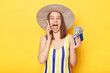 Unexpected vacation. Happy amazed woman on resort dressed in striped swimwear and straw hat posing isolated over yellow background holding passport and money talking phone boasting her vacation.