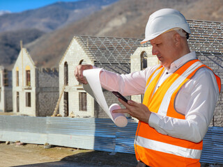 Poster - Man architect. Builder studies documentation. Worker near buildings under construction. Phone and piece of paper in hands builder. Man in helmet and reflective vest. Construction cottage settlement