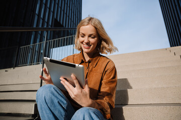 Smiling blonde woman using digital tablet while sitting on stairs outdoors