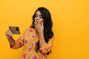 Wall Mural - Suprised indian woman covering her mouth while looking at mobile phone screen isolated over yellow background