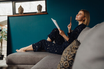 Wall Mural - Thoughtful woman writing down notes while sitting on sofa in her apartment