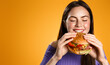 Portrait of young beautiful hungry woman eating burger. Isolated portrait of student with fast food over orange background. Diet concept
