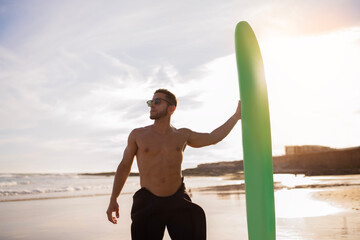 Wall Mural - Young Handsome Male Surfer With Surfboard Standing On Beach At Sunset