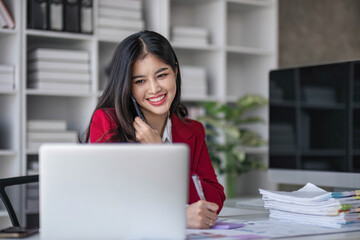 Young Asian businesswoman sit with her laptop computer happily talking on the phone with her customer explaining the detail in the office.