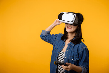 Poster - Young asian woman happy success wearing virtual reality headset game entertainment and holding joystick controller isolated on yellow background. Winner of cyber future video game concept.