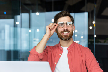 Wall Mural - Portrait of a successful and smiling young man with a beard and wearing a red shirt sitting in the office, working on a laptop and confidently looking to the side, holding his glasses with his hand.