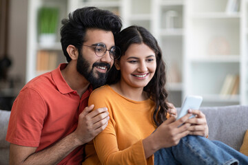 Wall Mural - Happy Young Indian Couple Watching Videos On Smartphone At Home Together