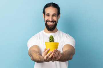 Portrait of handsome man bodybuilder with beard wearing white T-shirt holding out flower pot with cactus, looking at camera with toothy smile. Indoor studio shot isolated on blue background.