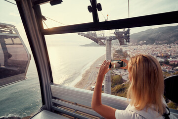 Wall Mural - Traveling by Turkey. Young tourist woman sitting in Alanya's cable car enjoying view taking photo on her smartphone.