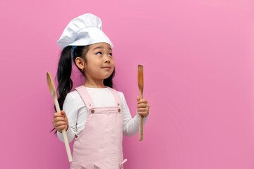 little asian girl in chef uniform holding kitchen items on pink isolated background and looking away at copy space