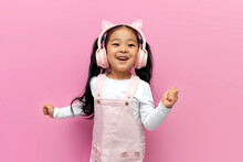 Little Asian Girl In Pink Children's Headphones Listens To Music And Dances On Pink Isolated Background, Korean Child