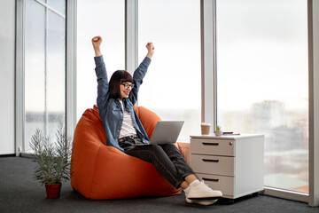 Excited caucasian lady rejoicing success while looking at laptop screen during work. Emotional freelancer in casual outfit holding remote gadget on knees and showing victory gesture with raised hands.