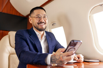 successful asian businessman in suit and glasses flies in private luxury jet and uses smartphone