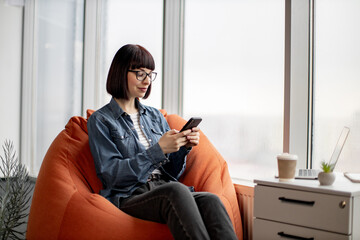 Beautiful young lady relaxing on comfortable office pouffe and holding smartphone in hands. Smiling female chatting in social networks, scrolling news line while resting near panoramic windows.