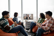 © sofiko14 - Focused diverse employees making video call on display while sitting with laptops and tablet on soft poufs in cozy workplace. Young self-employed workers taking part in online briefing in office.