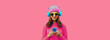 Portrait of stylish modern young woman listening to music with smartphone in wireless headphones wearing knitted sweater, hat on pink background