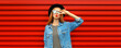 © rohappy - Portrait of stylish young woman model blowing her lips sending sweet air kiss wearing jean jacket, black round hat on red background