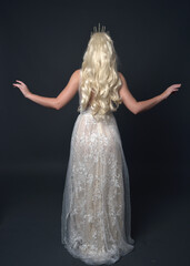 Wall Mural - Full length portrait of beautiful women with long blonde hair, wearing fantasy  princess crown and elegant white ball gown, standing pose with hand gesture. Isolated on dark grey studio background.