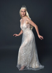 Wall Mural - Full length portrait of beautiful women with long blonde hair, wearing fantasy  princess crown and elegant white ball gown, standing pose with hand gesture. Isolated on dark grey studio background.