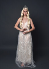 Wall Mural - Full length portrait of beautiful women with long blonde hair, wearing fantasy  princess crown and elegant white ball gown, standing pose with hand gesture. Isolated on dark grey studio background.