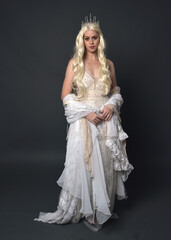 Wall Mural - Full length portrait of beautiful women with long blonde hair, wearing fantasy  princess crown and elegant white ball gown, standing pose with hand gesture. Isolated on dark grey studio background.