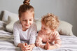 © sementsova321 - Sisters lying on bed in bedroom playing together relaxing in the morning talking having fun, caucasian kids at home in bedroom, wearing casual clothing.