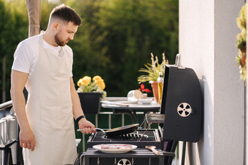 Handsome bearded man in apron frying potatoes on BBQ grill using frying carbon steel pan. 