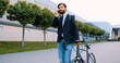 © serg - Portrait of good-tempered confident serious modern bearded man in stylish clothes which going down with his bike on the cobbled street and talking on smartphone