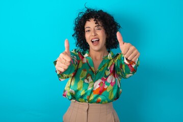 young arab woman wearing colorful shirt over blue background approving doing positive gesture with h