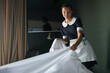 Young housekeeper or chambermaid in uniform changing blanket and other bedclothes on bed while preparing it for new guests of hotel
