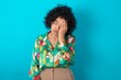 © Jihan - Tired overworked young arab woman wearing colorful shirt over blue background has sleepy expression, gloomy look, covers face with hand, has eyes shut, gasps from tiredness, fatigue after party
