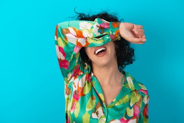 Sticker - young arab woman wearing colorful shirt over blue background covering eyes with arm smiling cheerful and funny. Blind concept.