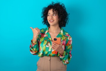 Sticker - young arab woman wearing colorful shirt over blue background using and texting with smartphone  pointing and showing with thumb up to the side with happy face smiling