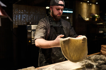 Young bearded chef in uniform holding dough flatbread over table sprinkled with flour while preparing pizza for restaurant guests