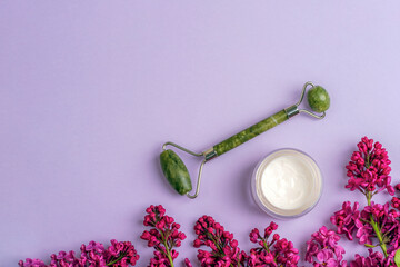 Wall Mural - Cream jar, jade face roller and lilac flowers on purple background. Natural cosmetics, skin care concept. Top view, flat lay, copy space