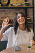 © Tatyana - Happy young dark-haired woman sitting in a cafe,waving her hand in a friendly way.