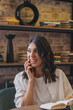 © Tatyana - Portrait of a smiling dark-haired young woman is talking on the phone with a book in her hands in a cafe.Remote work,business, freelance concept.