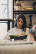 © Tatyana - A young attractive dark-haired woman is reading a book in a cafe and drinking green matcha tea.