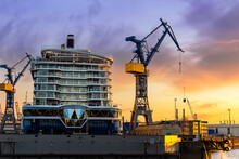 Scenic Back View Large Modern Luxury Cruise Ship Liner Under Construction Building Cranes At Dry Dock Shipyard In Hamburg Port Dramatic Sunset Sky Background. Big Vessel Manufactoring Site Industry