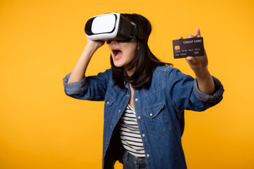 Poster - Young asian woman happy success wearing virtual reality headset game entertainment and holding credit card isolated on yellow background. Winner of cyber future video game concept.