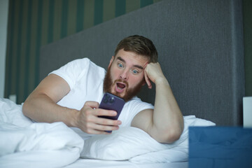 Canvas Print - Young bearded man is using cell mobile phone at home in bed in bedroom, looking at screen of smartphone 