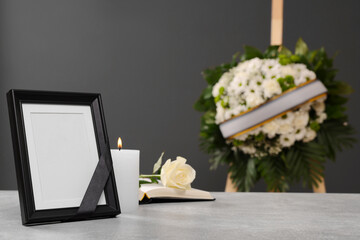 Wall Mural - Photo frame with black ribbon, rose, burning candle on light table and wreath of flowers near grey wall indoors, space for text. Funeral attributes