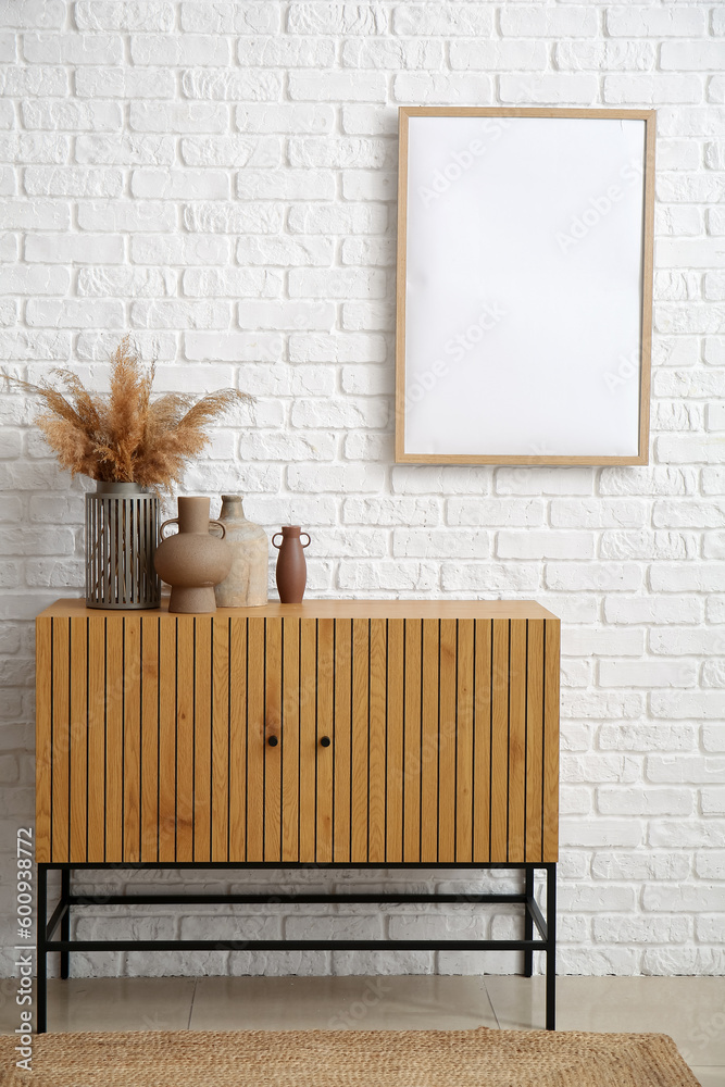 Wooden chest of drawers with vases near white brick wall in room